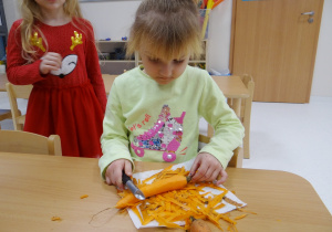 Ola siedzi przy stoliku i obiera marchewkę.